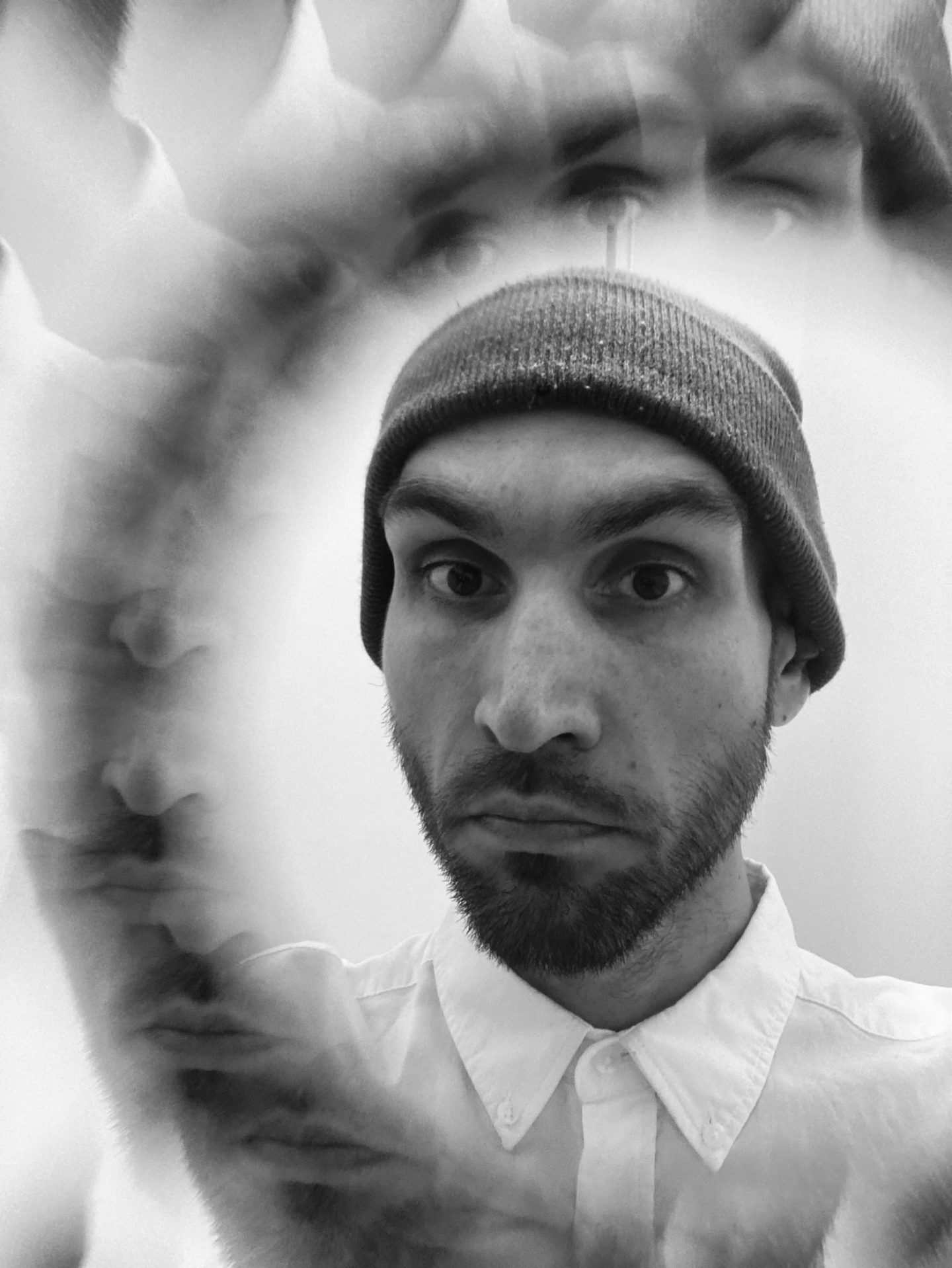 Black-and-white portrait of a person wearing a knitted beanie and white buttoned shirt, looking directly at the camera, surrounded by multiple blurred and overlapping images of the same face.
