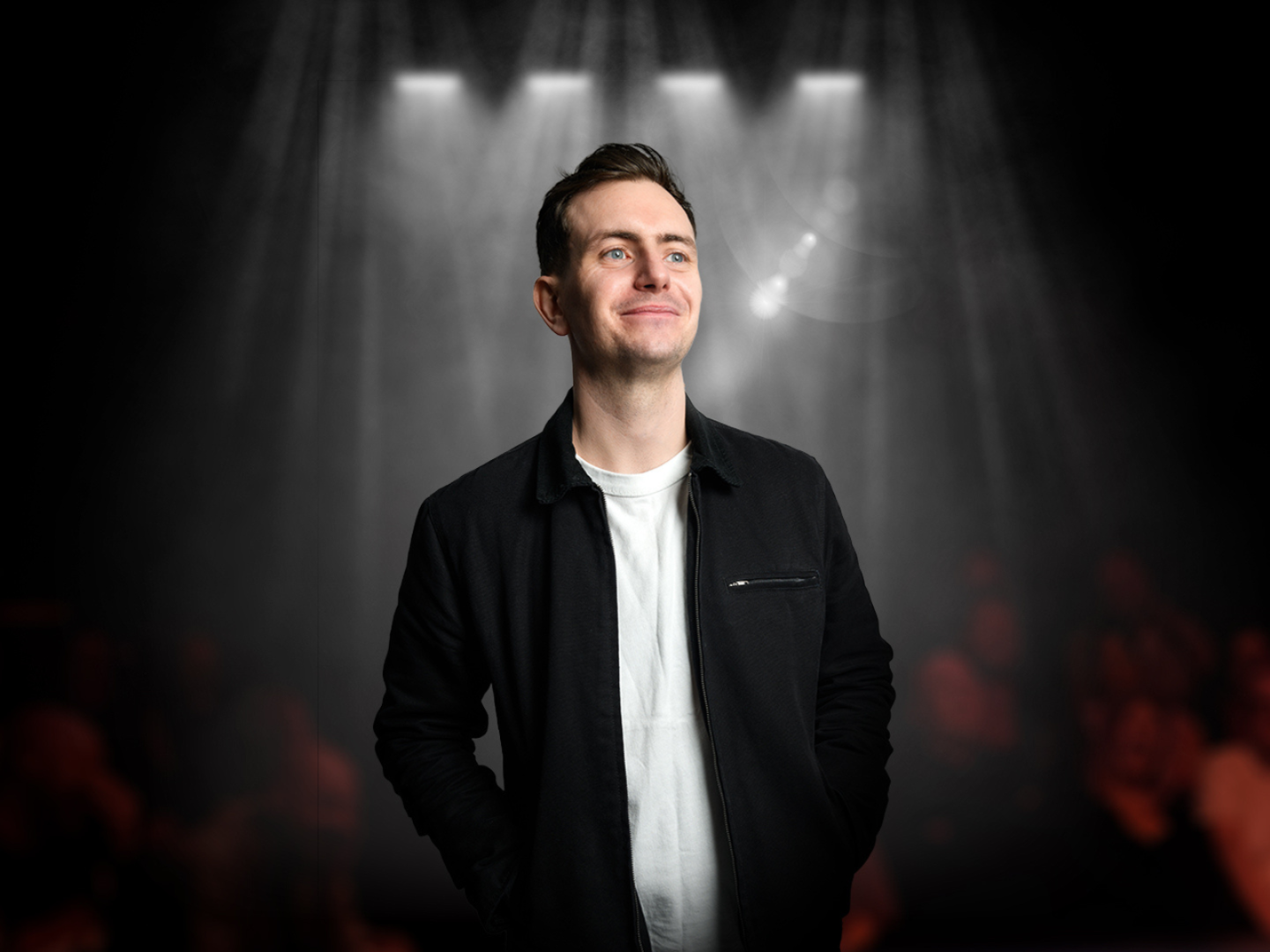Connor Burns standing on stage in front of spotlights and a dimly lit audience, wearing a black jacket over a white T-shirt.