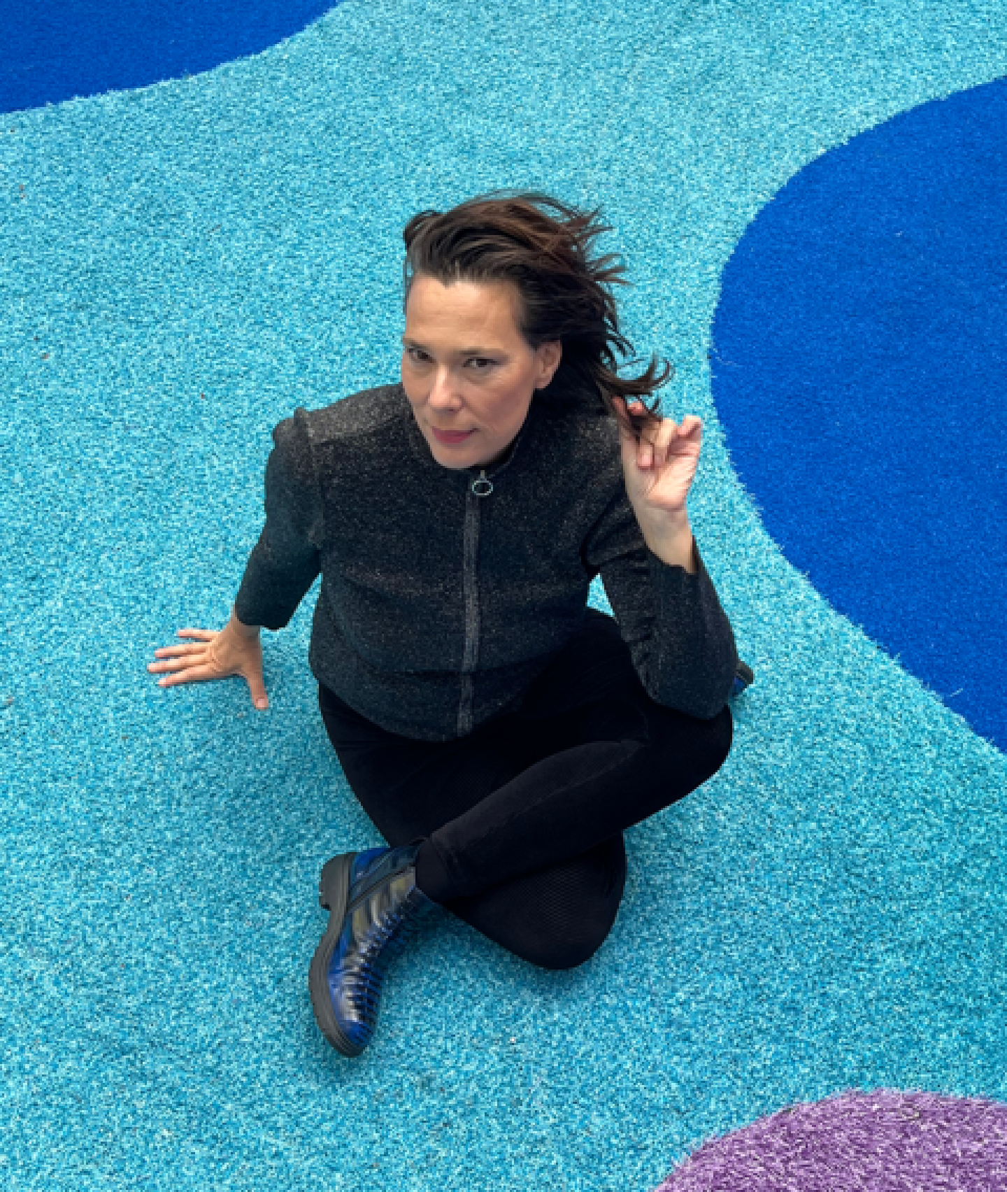 Person seated cross-legged on a bright turquoise and blue patterned surface, wearing a dark zip-up jacket, black trousers and dark shoes, photographed from above.