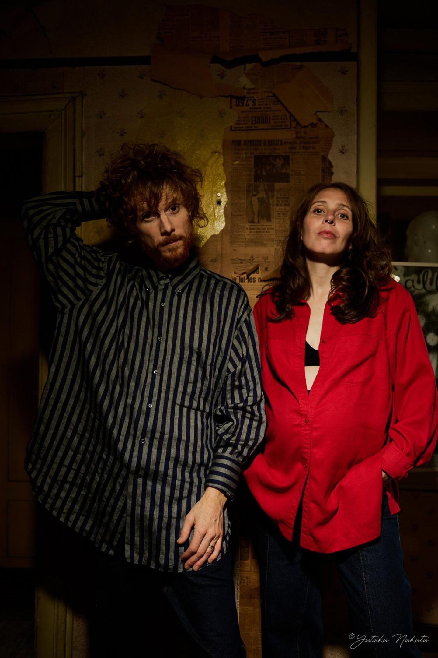 Two people standing side by side in a dimly lit interior. One wears a dark striped shirt and rests a hand behind their head; the other wears a bright red shirt. Torn newspaper pages cover part of the wall behind them.