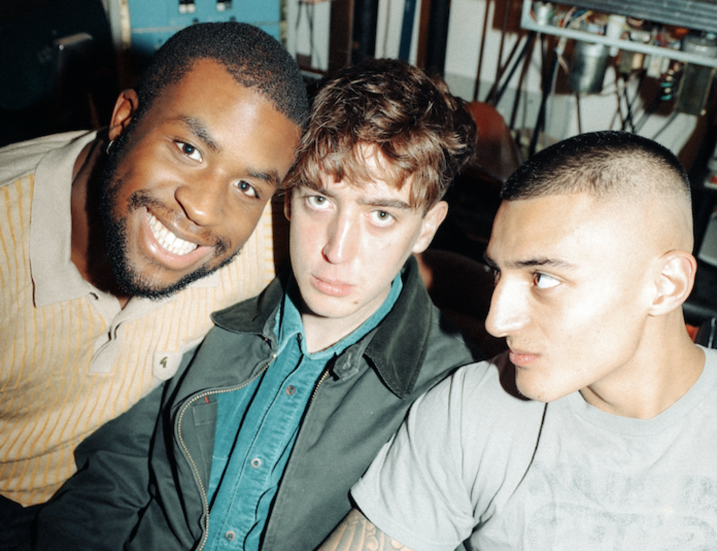 Three people, Knats, seated together indoors, photographed from above with flash lighting, with audio equipment and shelving visible in the background.