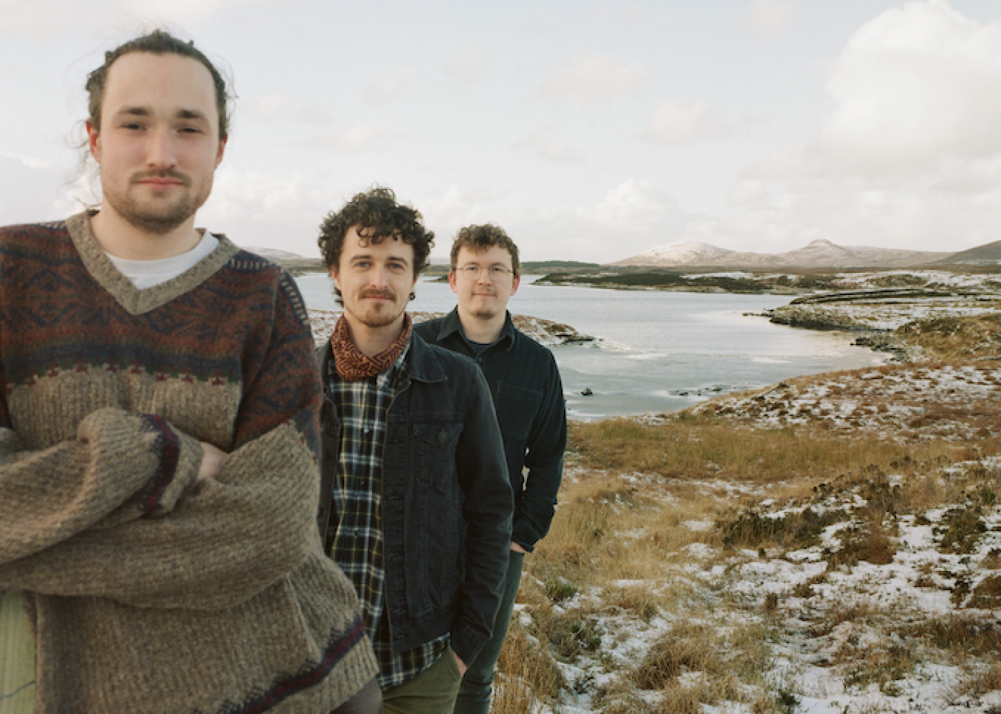 Three people standing outdoors near a coastal inlet, with snow-dusted hills and rugged grassland in the background.