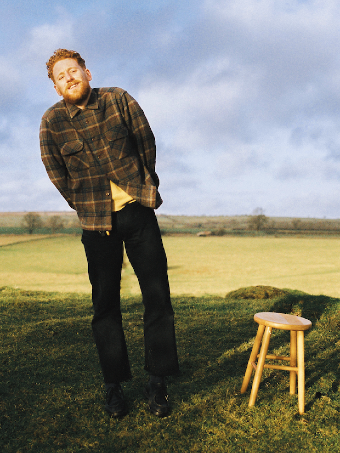 Willie J Healey standing in a grassy field beside a wooden stool under a cloudy sky.