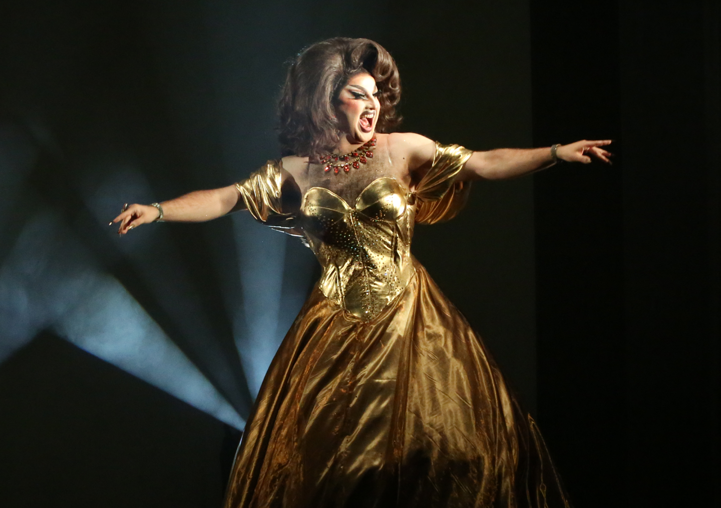 A drag performer in a glamorous gold satin gown, matching corset and statement red necklace poses dramatically on stage with arms outstretched, mouth open in an expressive performance. A spotlight shines through the dark, smoky background.
