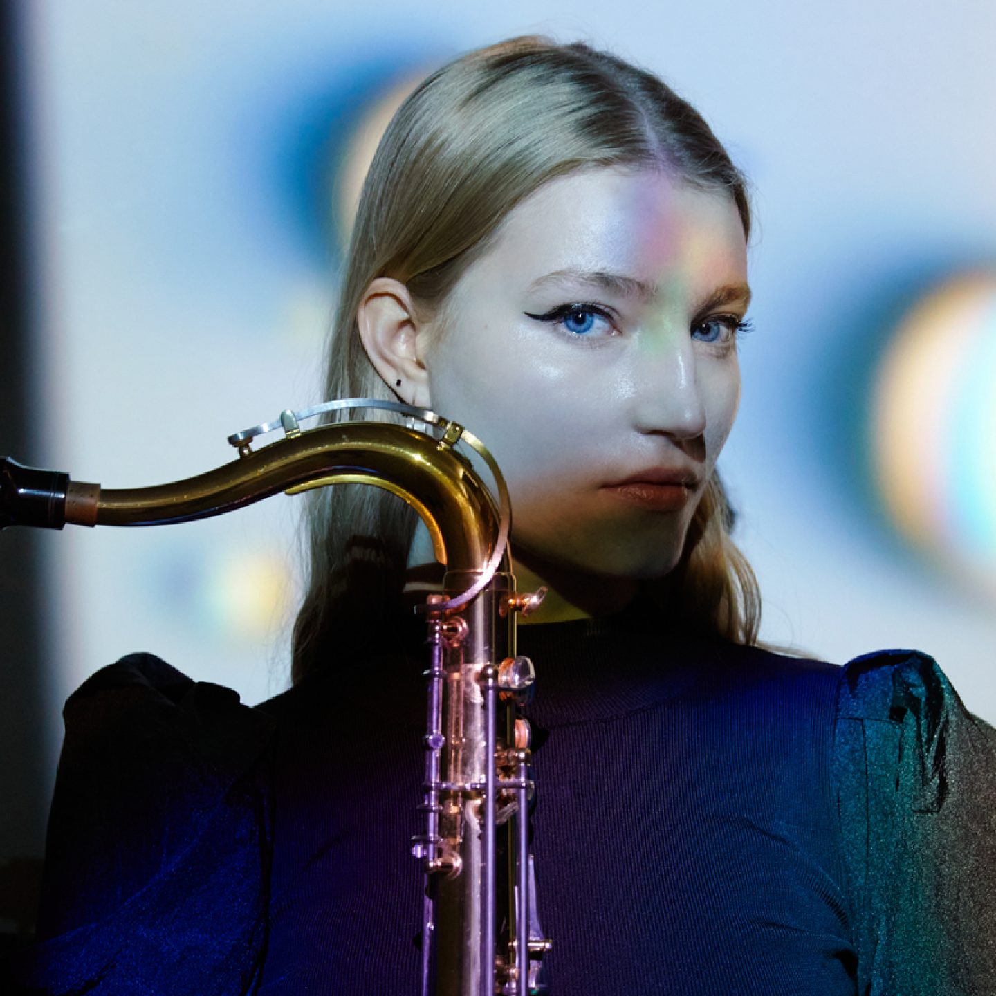 Close-up portrait of a Emma Rawicz holding a brass saxophone upright in front of their body. The person wears a dark, high-neck top and looks toward the camera. Soft blue, purple, and green lighting creates colorful reflections across the face, clothing, and instrument, while the background is blurred with circular light patterns.